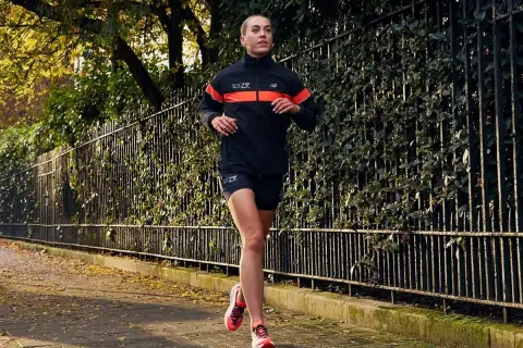 Kobieta w sportowym stroju biegnie po chodniku wśród jesiennych liści. Idealny strój, w czym biegać jesienią, zapewnia komfort i widoczność.