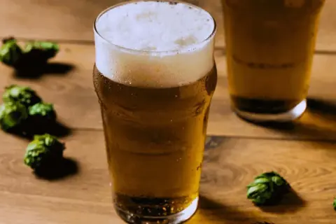 Un bicchiere di birra dorata con schiuma abbondante, circondato da luppoli verdi su un tavolo di legno. Perfetto per una festa della birra!