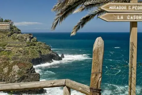 Cartel indicador en el sendero rambla de castro, señalizando Mirador S. Pedro y La Casona, con el mar azul de fondo.