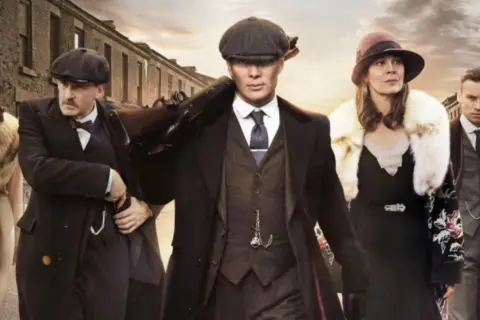 Gang w stylu Peaky Blinders, w tym Tommy Shelby z karabinem, idzie ulicą. Eleganckie płaszcze i kapelusze.