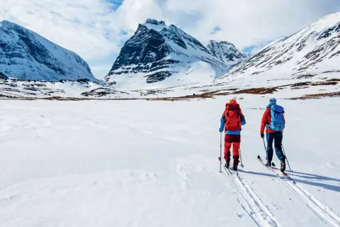 Två skidåkare med ryggsäckar på väg att bestiga berg i snöklädda fjäll under en molnig himmel.