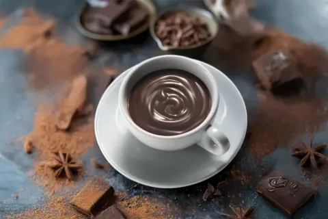 Una tazza cioccolata calda fumante, circondata da cacao in polvere, pezzi di cioccolato fondente e anice stellato.