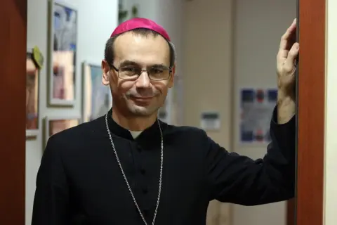 Uśmiechnięty, najmłodszy biskup w Polsce, w czarnej sutannie i z różowym zucchetto na głowie, opiera się o framugę drzwi.