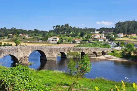 El ponte de Outeiro, un puente de piedra con varios arcos, cruza un río azul. En la orilla opuesta, casas con tejados rojos se asientan en una colina verde.
