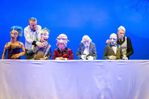 Puppenspieler dirigieren ihre Figuren auf der Bühne im Theater Remscheid. Eine bunte Truppe von Marionetten sitzt hinter einem weißen Tuch.