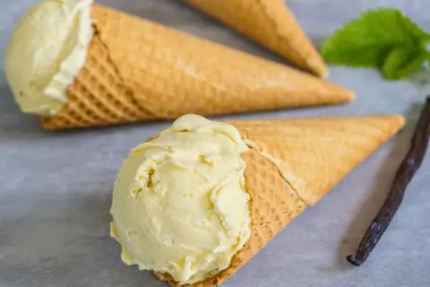 Drei Waffelkegel mit cremigem Vanilleeis, daneben eine Vanilleschote und ein Minzblatt.