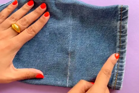 Main d'une femme avec un ongle rouge vif et une bague dorée, ajustant l'ourlet d'un jean.