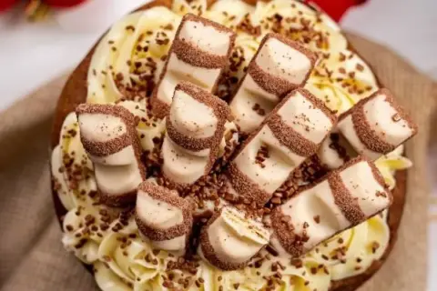 Tarta decorada con crema amarilla y trozos de tarta kinder bueno, espolvoreada con virutas de chocolate.