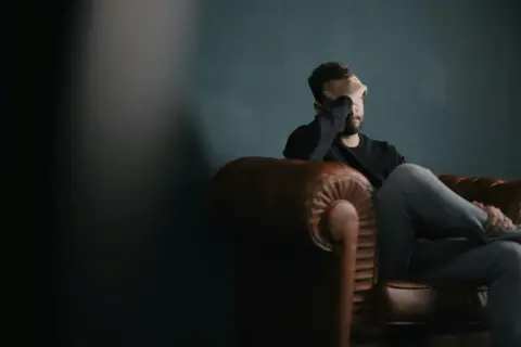 Mężczyzna siedzi na skórzanej kanapie, zasłaniając twarz dłonią. W jego postawie widać przygnębienie i myśli rezygnacyjne.