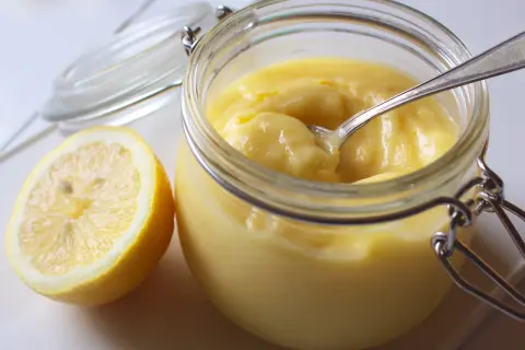 En burk med krämig lemon curd och en halv citron bredvid. En sked vilar i burken, redo att skopa upp den syrliga godsaken.