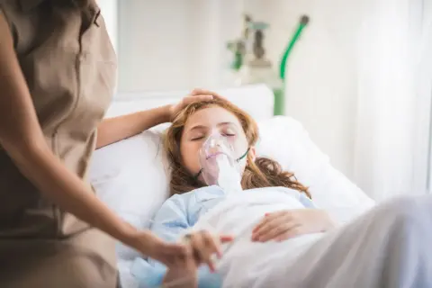 Dziewczynka w szpitalnym łóżku z maską tlenową, walcząca z hipoksją. Opiekunka czule kładzie dłoń na jej głowie.