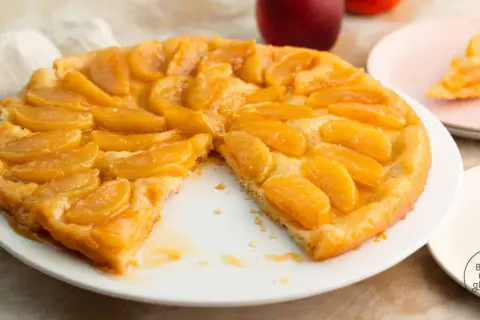 Eine goldbraune Tarte Tatin mit karamellisierten Apfelspalten auf einem weißen Teller. Ein Stück fehlt, was Appetit macht.