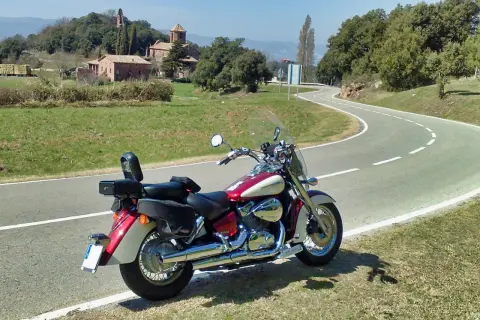 Honda VT 750 C Shadow 2007 ficha técnica. Moto custom roja y crema aparcada en una carretera rural con un paisaje de colinas y edificios antiguos.