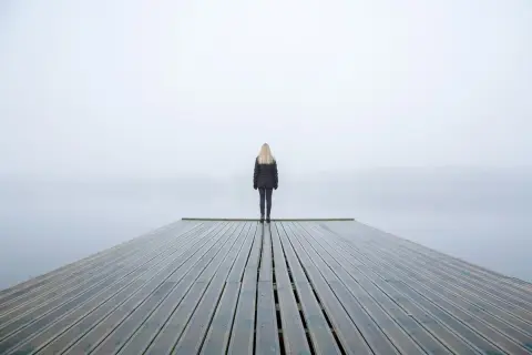 Una persona fredda, con la psicologia che la isola, sta in piedi su un molo di legno in una giornata nebbiosa.