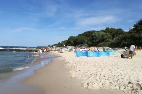 Sianożęty atrakcje: plaża pełna ludzi, falochrony w morzu, zielony las w tle.