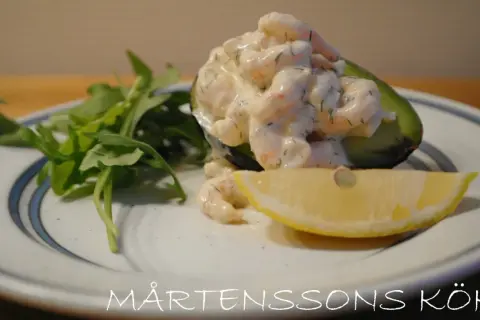 Avokadohalva fylld med räkor i en krämig sås, liknande guacamole crème fraiche, med en citronskiva och gröna blad.