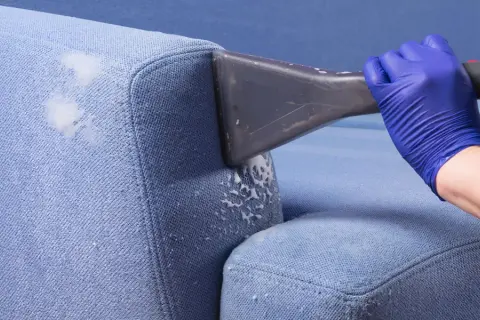 Blaue Polster reinigen mit Rasierschaum. Ein Handschuh hält ein Reinigungsgerät, das Schaum auf den Stoff sprüht.