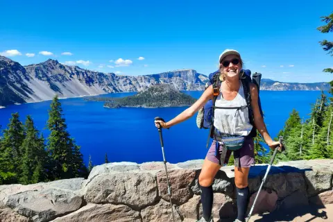 Uśmiechnięta turystka z plecakiem i kijkami, podziwiająca błękitne jezioro. To widok z trasy Pacific Crest Trail, pokazujący, ile się idzie przez takie cuda natury.
