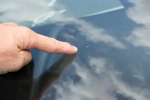 Dedo señala un pequeño impacto en el parabrisas, un cristal manchado por dentro por la suciedad y el reflejo del cielo nublado.