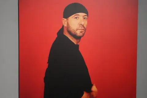 Retrato de Manuel Piña con gorro y camiseta negra, sobre fondo rojo vibrante.