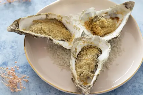 Trois huitres au four, garnies de chapelure dorée, reposent sur un lit de gros sel sur une assiette rose.