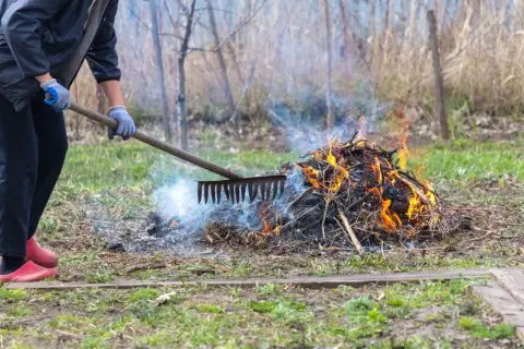 Osoba w rękawiczkach grabi ognisko na posesji, z którego unosi się dym i płomienie.