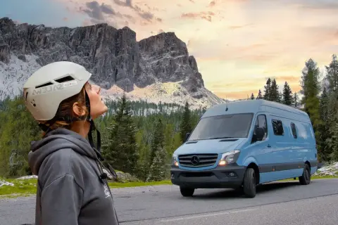 Mujer con casco mira las montañas. A su lado, un **autobús camperizado** azul espera para la aventura.