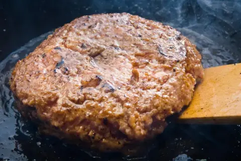 Jugosa carne dorándose en sartén, parte de como hacer hamburguesas caseras. Humo y burbujas de grasa.