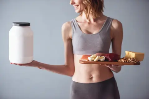 Femme souriante tenant un pot de protéines et une assiette avec viande, œufs, fromage et noix. Idéal pour le calcul protéine par jour femme.