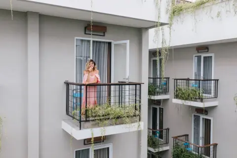 Kobieta w różowej sukience stoi na balkonie, opierając się o balustradę. Wokół rosną zielone rośliny, tworząc naturalną zasłonę na balkon.