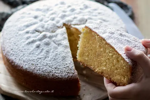 Una fetta di torta margherita soffice, cosparsa di zucchero a velo, viene sollevata da una mano.