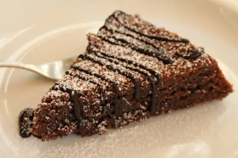 Una fetta di torta caprese al cioccolato, cosparsa di zucchero a velo e con una colatura di cioccolato fondente.