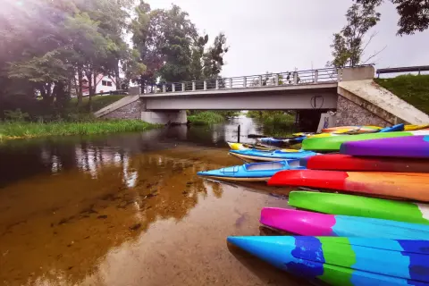 Kolorowe kajaki czekają nad rzeką, idealne na spływ kajakowy Mazury gdzie najlepiej. Most i zieleń tworzą malowniczy krajobraz.