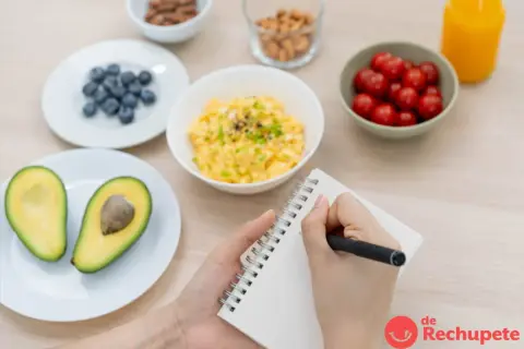 Manos escribiendo en un cuaderno, rodeadas de ingredientes para crear ejemplos de menú: aguacate, arándanos, huevos revueltos, tomates cherry, frutos secos y jugo.
