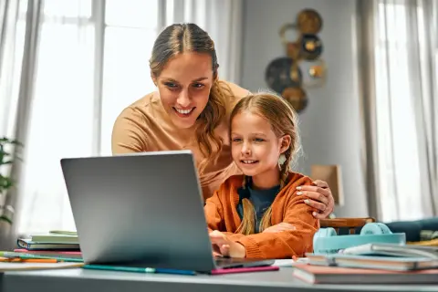 Uśmiechnięta mama wspiera córkę w nauce przy laptopie. To piękny przykład opieki na dziecko, wspólnego spędzania czasu i wspierania rozwoju.