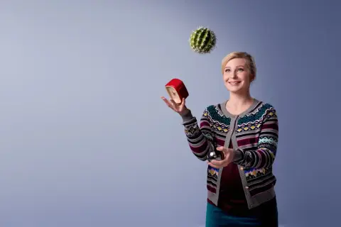 Kvinna jonglerar med en kaktus, en röd väckarklocka och en metallkula. Hon ser glad ut, som om hon klarar allt. Osa 2015.