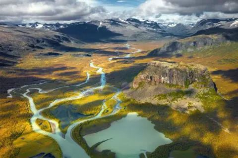 Weites Tal mit Fluss und Berg in Schwedens Nationalparks. Herbstliche Farben und dramatische Wolken über der Landschaft.