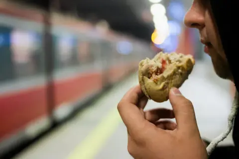 Osoba w kapturze je bułkę na tle pociągu TLK.
