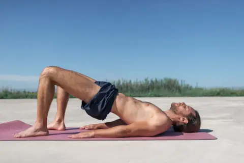 Homme pratiquant l'exercice de Kegel sur un tapis rose, le corps surélevé, les genoux pliés, sous un ciel bleu clair.