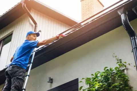 Man on ladder cleaning gutters. This task should be done at least twice a year to prevent clogs.