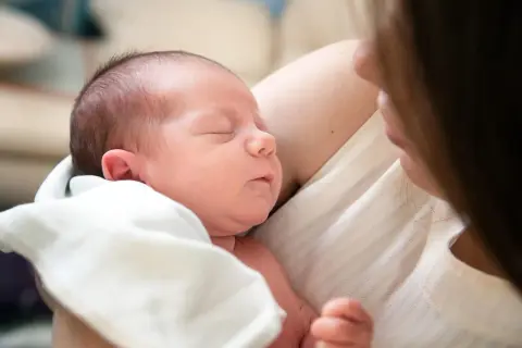 Śpiące, 3-tygodniowe dziecko wtulone w ramiona mamy. Delikatne światło podkreśla jego maleńkie rysy.