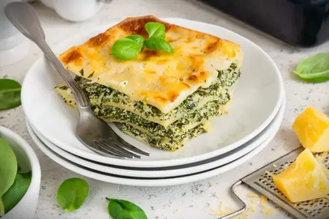 Ein Stück Spinatlasagne mit Käse überbacken, garniert mit Basilikum, auf einem Teller mit einer Gabel.