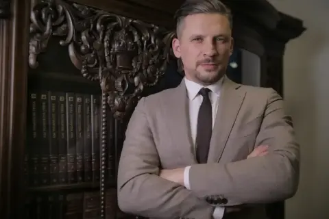 Mężczyzna w garniturze stoi przed regałem z książkami, ręce skrzyżowane na piersi.