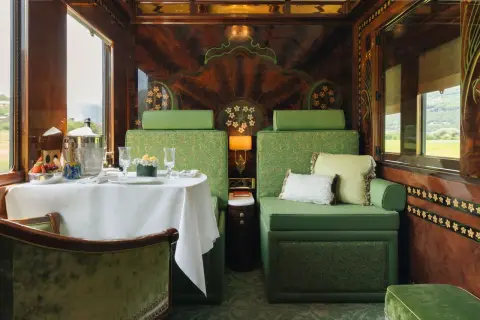 Luxuriöser Speisewagen im Orient Express: Ein gedeckter Tisch mit Obst und Champagner, grüne Sitzmöbel und kunstvolle Holzverzierungen.