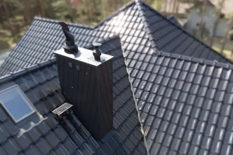 Czarny, nowoczesny komin na dachu z dachówki ceramicznej. Obok okno dachowe i stopień kominiarski.