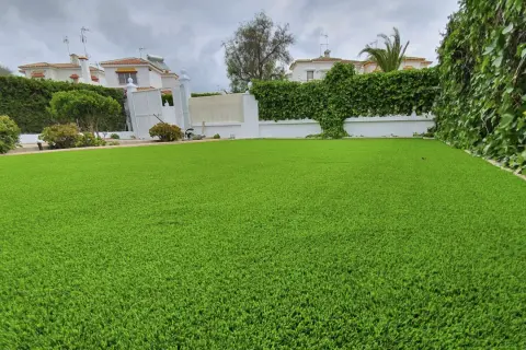 Un jardín con césped artificial impecable, delimitado por vegetación exuberante y casas blancas al fondo.