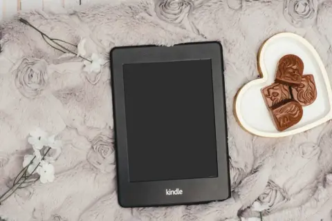 Un Kindle Vella posé sur une couverture douce, avec des chocolats en forme de cœur et des fleurs blanches.