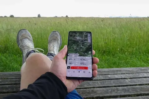 Person sitzt auf einer Holzbank, hält ein Smartphone mit einer guten Wander-App, die eine 6,74 km lange Tour mit 16 hm Anstieg und 672 hm Seehöhe anzeigt.