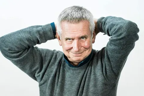 Uśmiechnięty mężczyzna z siwymi włosami, który trzyma ręce za głową. To on jest tym, kto pisze teksty żartów i anegdot, które opowiada prowadzący show.