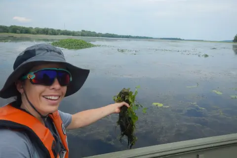 Une femme en gilet de sauvetage tient une plante aquatique, peut-être une châtaigne d'eau, dans un lac.